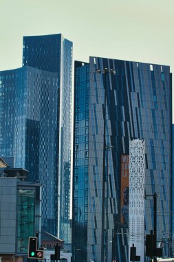 Görüntüde birden fazla modern, mavi tonlu gökdelenler ve Manchester, İngiltere 'de solgun bir gökyüzünün altında kentsel bir ortamda süs kulesi var..