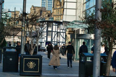 Manchester, İngiltere 'de eski ve modern binaların, sokak mobilyalarının ve sessiz renklerin yer aldığı kaldırımlı bir kaldırımda yürüyen yayaların olduğu bir sokak sahnesi..