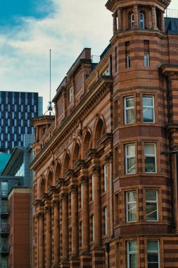 Görüntü, arka planda modern bir bina ve Manchester, İngiltere 'de bulutlu bir gökyüzü ile vurgulanmış, sütunları ve kemerleri olan kahverengi tuğlalı bir bina gösteriyor..