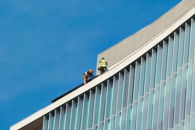 Manchester, İngiltere 'de açık mavi gökyüzüne bakan cam cepheli modern bir binanın çatısında güvenlik teçhizatlı iki işçi..