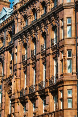 Detaylı bir mimari bakış açısında, Manchester, İngiltere 'de kahverengi taş bina cephesi, kemerli camlar, süslü demir balkonlar ve ağır gölgeli detaylar yer alıyor..