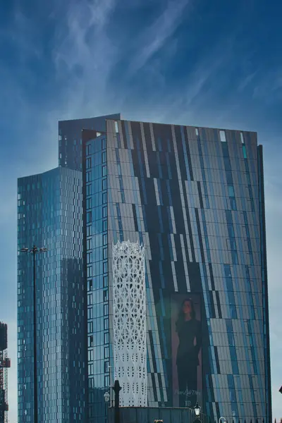 Mavi ve gri cepheli uzun modern binalar ince bulutlu mavi gökyüzüne karşı. Manchester, İngiltere 'de bir binanın temelinde beyaz bir dekoratif yapı bulunmaktadır..
