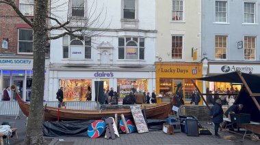 Açık hava manzarası, York, İngiltere 'de insanlarla dolu bir caddede çeşitli dükkanların önünde ahşap bir tekne, kalkanlar ve market tezgahları gösteriyor..