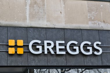 Greggs mağaza tabelası. Beyaz harfler ve turuncu kareler koyu gri bir zemin üzerinde, Liverpool, İngiltere 'de daha açık renkli taş bir duvar var..