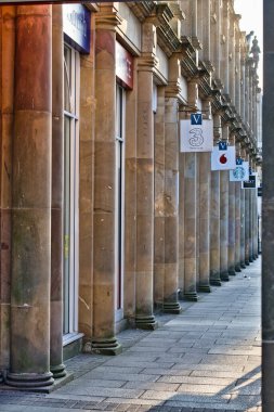 Bir şehir caddesini güneş ışığıyla aydınlatan, vitrinlerle sıralanmış sütunlar. Pankartlar ve pankartlar Harrogate, Yorkshire, İngiltere 'deki mimari detayların altında asılı..
