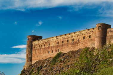 Tepenin üzerindeki tuğla şatoda kuleler, bir saat ve İngiltere 'deki Bamburgh Kalesi' nde dağınık bulutlu mavi bir gökyüzünün altında heybetli bir duvar var..