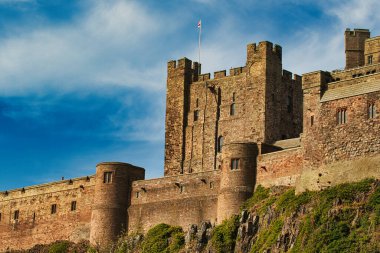 İngiltere 'nin Bamburgh Şatosu' nda yeşilliklerle kaplı kayalık bir çıkıntıda mavi gökyüzüne karşı taretleri olan büyük, taştan bir şatonun alçak açılı görüntüsü..