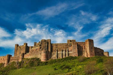 Çimenli bir tepedeki taş bir kalenin geniş bir görüntüsü, Birleşik Krallık Bamburgh Kalesi 'nde dağınık beyaz bulutlarla mavi bir gökyüzüne karşı kurulmuş..