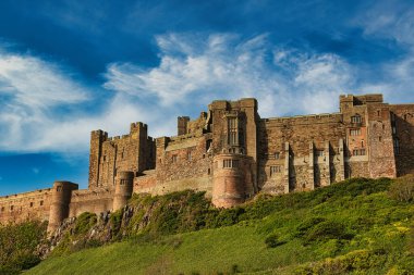 İnce bulutlu mavi bir gökyüzünün altındaki yeşil bir tepenin üzerinde büyük bir taş kale bulunur. Kale, Birleşik Krallık 'taki Bamburgh Kalesi' nin baskın unsurudur..