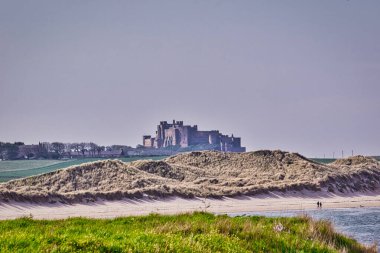 Tepenin tepesindeki bir kalenin manzara görüntüsü, çimenli kum tepeleri, kumlu bir sahil ve su. İnsanlar İngiltere 'deki Bamburgh Kalesi' nde kıyı şeridi boyunca yürüyorlar..