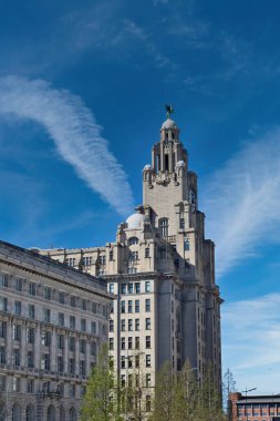 Görüntü, Liver Binası 'nı, İngiltere' nin Liverpool kentindeki daha küçük bitişik bir binanın yanında, mavi bir gökyüzünün altında, çizgili bulutlarla gösteriyor..