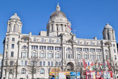 Görüntü Liverpool 'daki Kraliyet Karaciğer Binası' nı gösteriyor. Arkaplanı açık mavi gökyüzü olan büyük, süslü bir bina. Tur otobüsleri ön planda..