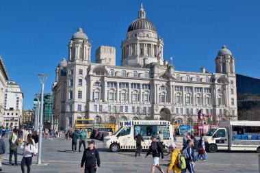 Görüntü, İngiltere 'nin Liverpool kentindeki Kraliyet Karaciğer Binası' nı gösteriyor. Önünde açık mavi gökyüzünün altında insanlar ve yemek kamyonları var..