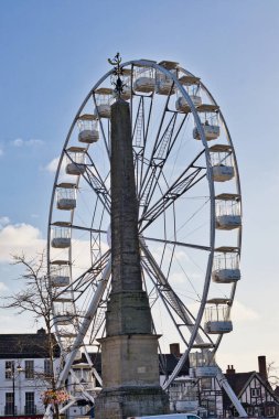Bir dönme dolap, Ripon, Yorkshire, İngiltere 'de arka planda dağınık bulutlar ve binalar ile parlak mavi bir gökyüzüne karşı duran uzun bir taş anıtın arkasında duruyor..