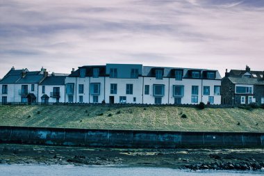 Çimenli bir bankanın tepesinde, beton bir duvar ve kayalık bir kıyı şeridinin üzerinde, Seahouses Harbour, İngiltere 'de bulutlu bir gökyüzünün altında, sıra sıra evler bulunuyor.