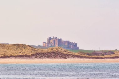 Ön planda kum tepeleri, arka planda plaj ve kale, soluk gökyüzünün altında Bamburgh Şatosu, İngiltere.