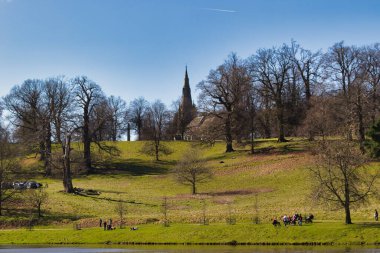Tepede bir kilise kulesi, çıplak ağaçlar, insanlar ve İngiltere 'deki Studley Royal Park' ta açık mavi gökyüzünün altında bir su kütlesi..
