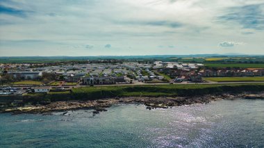 İngiltere 'nin deniz evleri köyünde parçalı bulutlu bir gökyüzü altında seyyar ev parkı, okyanus ve tarlaları olan bir sahil kasabasının havadan görünüşü..