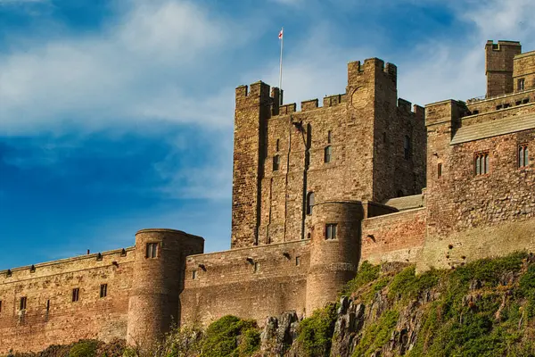 İngiltere 'nin Bamburgh Şatosu' nda yeşilliklerle kaplı kayalık bir çıkıntıda mavi gökyüzüne karşı taretleri olan büyük, taştan bir şatonun alçak açılı görüntüsü..