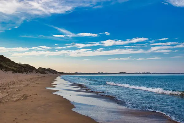 Kumlu bir sahil manzarası, kumullar, okyanus dalgaları ve dağınık bulutlu mavi bir gökyüzü..