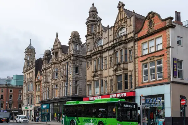 Sokak manzaralı gösterişli, çok katlı binaların önünde yeşil bir otobüs var. Yayalar ve dükkanlar Leeds, İngiltere 'de caddede kuyruk oluşturuyorlar..