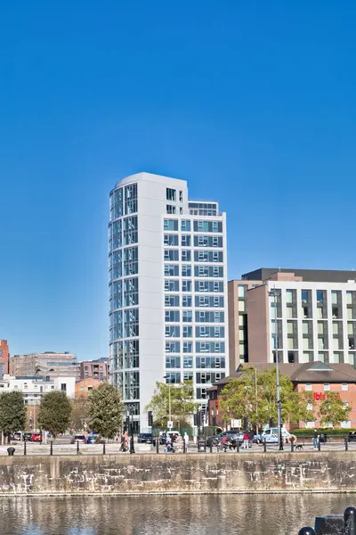 Mavi pencereli uzun beyaz bir bina, üzerinde nehir önplanı ve mavi gökyüzü, diğer binalar ve İngiltere 'nin Liverpool kentindeki insanlarla merkezlidir..