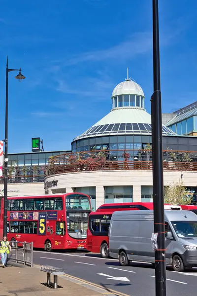 Kırmızı otobüslerin göz hizasında görüntüsü, gri bir minibüs ve Wimbledon, Londra, İngiltere 'de açık mavi gökyüzüne karşı cam kubbeli çok katlı bir bina..
