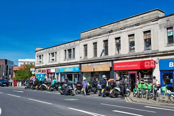 Park halindeki motosikletleri, aralarında Greggs ve British Heart Foundation 'ın da bulunduğu dükkanların önünde, Wimbledon, Londra' da açık mavi bir gökyüzünün altında gösteriyor..