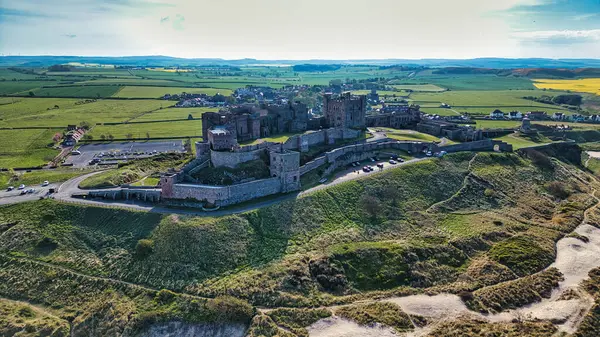 Çimenli bir tepenin üstündeki büyük bir taş şatonun havadan görünüşü, tarlalara bakan bir tepe ve parlak bir gökyüzünün altındaki küçük bir köy..