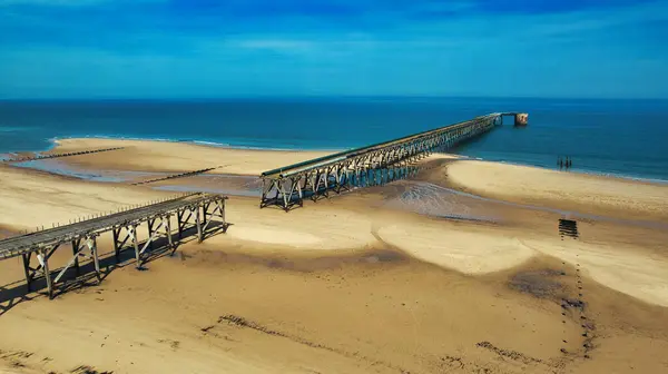 İngiltere 'nin Steetley İskelesi' ndeki mavi gökyüzünün altında kumlu bir plajdan okyanusa uzanan iki harap ahşap iskele görüntüsü..
