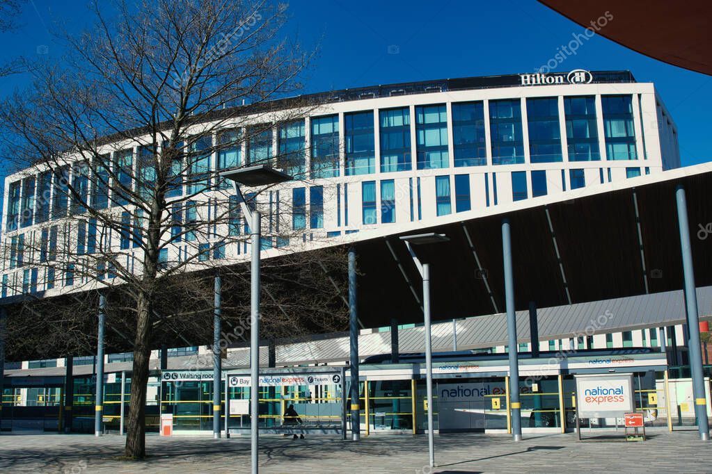 Exterior shot of a Hilton Hotel behind a bus depot, with a bare tree on the left and a bright blue sky in Liverpool, UK.