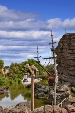 Etrafı kayalar, yeşillikler ve Fransa 'nın Disneyland şehrinde parçalı bulutlu bir gökyüzü ile çevrili bir göle yanaşmış bir korsan gemisinin manzarası..