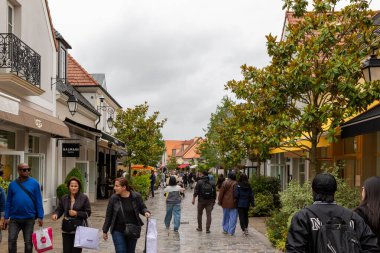 Fransa 'nın La Vallee Köyü' nde bulutlu bir gökyüzünün altında ağaçlarla ve dükkanlarla dolu bir kaldırım taşından sokakta yürüyen insanların görüntüsü..