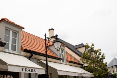 Turuncu kiremit çatılı ticari binaların dış görünüşü, 'PRADA' tabelası olan tenteler ve Fransa 'nın La Vallee Köyü' nde bulutlu bir gökyüzüne karşı siyah bir sokak lambası..