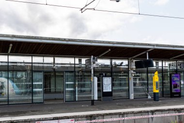 Cam pencereli tren istasyonu platformu, tepe çizgileri, tabela ve bulutlu bir gökyüzünün altında sarı bir makine..