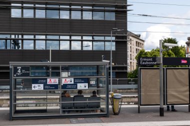 Train station platform with a shelter, information signs, and a modern building in the background.