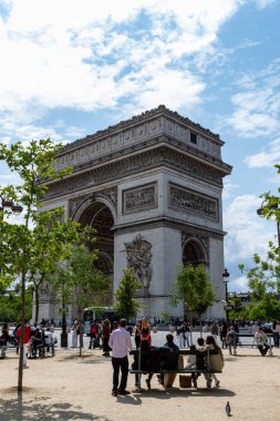 The Arc de Triomphe is viewed from a park-like setting with trees and people. Blue sky and clouds provide a backdrop.