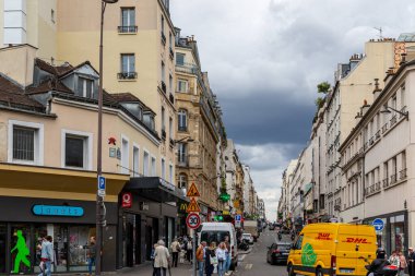 Street view in Paris with buildings, shops, traffic, and pedestrians under a cloudy sky. Predominantly beige, yellow and white building colors.