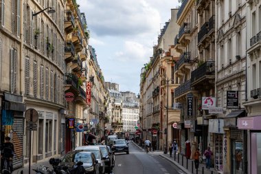 Paris, Fransa 'daki sokak manzarasında bulutlu bir gökyüzü altında binalar, dükkanlar, park edilmiş arabalar ve yayalar yer alıyor.