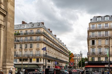 Paris 'in sokak manzarası, çok katlı binalar, bazıları balkonlu, bulutlu bir gökyüzü altında. Sokak seviyesindeki işletmeler görülebilir.