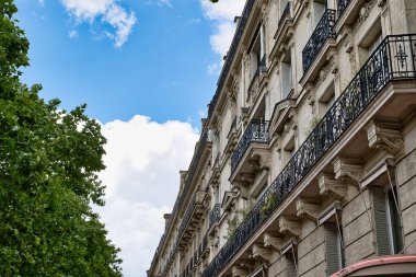 Bulutlu bir gökyüzüne karşı balkonlu bir binanın alçak açılı görüntüsü ve sol köşede ağaçlar..