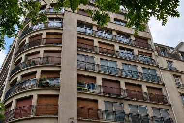 Üst ve sol köşesindeki ağaç yapraklarıyla çerçevelenmiş, balkonlu ve nötr tonlu çok katlı bir binanın alçak açılı görüntüsü..