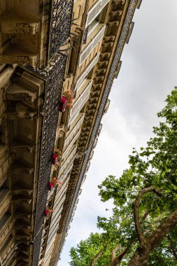 Low-angle view of a building with ornate facade, white shutters, red flower boxes, and wrought-iron balconies, framed by trees and a cloudy sky.