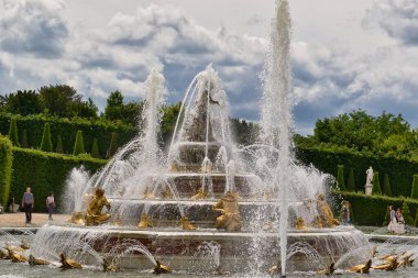 Katmanlı havzalar, altın heykeller ve gökyüzüne yükselen su jetleriyle süslü bir çeşme, manikürlü çitlere karşı kurulmuş ve Paris Versailles Gardens 'da bulutlu bir gökyüzü.