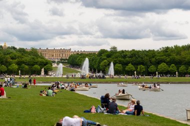 Versailles Gardens sahnesinde çimenlerde dinlenen insanlar, suda kayıklar, fıskiyeler ve bulutlu bir gökyüzünün altında saray.