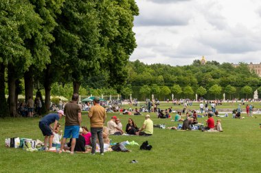 Güneşli bir park sahnesinde büyük yeşil bir çimenlikte piknik yapan insanlar görülüyor. Çevreyi çevreleyen ağaçlar, Versailles Gardens, Paris 'teki binalarla çevrili..
