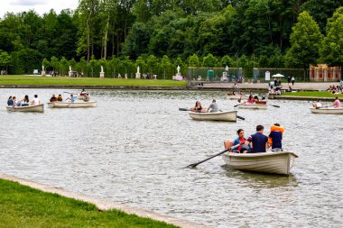 Gölde kayıkla gezen insanlar, sahilde yeşil ağaçlar ve heykeller görülebiliyor. Versailles Gardens, Paris 'te bulutlu bir gökyüzü.