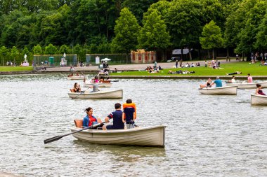 Gölde içinde insanlar olan birkaç kayık; insanlar Paris 'teki Versailles Gardens' ın arka planındaki çimenli sahilde dinleniyor..