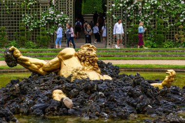 Çeşmedeki altın heykel, etrafı siyah kayalarla çevrili. İnsanlar arka planda yürüyor, Paris 'teki Versailles Gardens' ta kemerli, çiçekli bir çardak tarafından çerçevelenmiş..