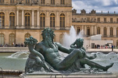 Versay 'daki bir çeşmede bronz bir Neptün heykeli. Versailles Sarayı arka planda bulutlu bir gökyüzünün altında..